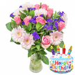 Bathed in soft, even light against a clean white backdrop, this celebratory arrangement reads as a bright, tactile gift: a lush bouquet of freshly cut blooms set in a clear glass vase with green stems and water visible at the base, paired with a playful cake-shaped foil balloon. The flower composition centers on fully opened soft-pink roses, their velvety petals unfurling from pale blush to a warmer, more saturated pink at the hearts, while generous pale-pink Gerbera daisies-each with layered, ray-like petals and buttercream-yellow centers-form cheerful focal points that feel almost like smiling faces. Woven between the larger blossoms are clusters of vivid purple filler flowers, likely statice or limonium, whose crisp, papery texture gives depth and a jewel-like contrast to the pinks. Small white spray blooms add flecks of brightness while glossy, deep-green foliage frames and supports the whole, giving the bouquet a natural, freshly gathered look.

To the right, a white, cake-shaped Mylar balloon stands as a festive partner: a frosted surface trimmed with a wavy blue icing border, scattered multi-colored sprinkles in red, yellow, blue and green, and four striped candles-purple, red, orange and blue-each topped with a tiny yellow flame. The words Happy Birthday appear across the front in playful, multi-colored type and a small KALEIDOSCOPE logo sits at the balloon's base. There is a gentle, sweet fragrance suggested by the roses and greens, and the overall impression is joyous and affectionate-an ideal birthday gesture from a florist who values texture, colour and freshness.