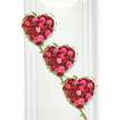 In this scene, three heart-shaped floral arrangements drift gracefully downward in a diagonal line, each one completely filled with roses in sumptuous reds and tender pinks. The hearts are tightly composed, every bloom positioned so that petals blend into petals, forming a rich, continuous layer of colour and softness. The deepest crimson roses lend a sense of intensity and enduring love, while vivid pinks and delicate blush tones lighten the design, echoing memories of joy and affection shared. Some roses stand wide open, showing their full spiral of petals, others are caught mid-bloom, and a few remain as rounded buds, suggesting that love and remembrance continue to unfold even as we say goodbye. Around the edge of each heart, slender strands of ivy twist and trail, their small, glossy green leaves providing a natural outline and linking the hearts into a single, flowing tribute. These gentle green lines bring a sense of the outdoors, reminiscent of the leafy calm around Grove Park, its riverside paths and tree-shaded streets where families walk and reflect. The hearts rest against a perfectly white background that looks like a smooth, pristine panel or wall, similar to what you might find in a local chapel of rest or a dedicated tribute area in a Grove Park funeral venue. Soft, balanced lighting reveals the depth and variation in every petal, while faint shadows on one side give shape and dimension to the forms. The overall feeling is one of quiet romance mixed with solemn respect, making this arrangement especially appropriate as a funeral tribute, remembrance display, or anniversary of loss, where the message of enduring love is spoken gently through the language of roses and ivy.