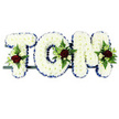 A carefully crafted floral name tribute spells out TOM in three capital letters, designed in the understated, traditional style often chosen for funerals and memorials in Grove Park. Each letter is completely filled with pristine white chrysanthemums, the small, rounded heads packed so tightly that they form a dense, cushion-like surface, suggesting softness and serenity. Around the outer edge of every letter, a royal blue satin ribbon is carefully pleated and pinned, tracing the contours of the T, the circular O, and the angled M. This ribbon border clearly defines each shape and adds a refined, formal note that frames the white blooms beautifully. Positioned at thoughtful points across the letters are small but expressive floral sprays: rich red roses with velvety petals serve as the central feature, surrounded by tiny white and cream spray roses and graceful white blossoms similar to freesia, which introduce a hint of light fragrance and delicacy. These flowers rest in a collar of mixed greenery, with feathery ferns and glossy leaves providing texture, depth, and a touch of natural movement as they gently fan out around the blooms. The letters sit on a slim dark support bar, aligned side by side in a straight line, as they would be on display at a chapel, crematorium, or graveside service nearby. The plain white background keeps the focus firmly on the tribute itself, making the contrast between the white chrysanthemum base, the blue ribbon edging, and the red rose accents especially striking. Overall, the design conveys a quiet sense of respect, love, and remembrance for someone named Tom, offering a comforting visual focus for family and friends gathering in Grove Park to say their final farewells.