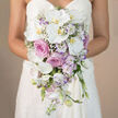 A bride in a smooth white sweetheart gown is shown from the waist down, the soft curve of the dress acting as a backdrop for an extravagant cascading bouquet that tumbles from her hands like a floral waterfall. The background is a neutral beige blur, allowing the detail of each flower to shine. Dominating the composition are sweeping white Phalaenopsis orchids, their petals broad, silky, and almost luminous, each bloom featuring a small yellow-orange centre that glows warmly amid the cooler tones. Tucked into and around the orchids are lush pink roses and full-bodied garden roses in a dusty lavender-pink shade, their layered petals giving a sense of romance and quiet luxury. Among them, white lisianthus with subtle purple-edged petals and slender stalks of light lavender stock introduce further nuance, both in colour and in the bouquet's structure. Wisps of white baby's breath thread delicately through the arrangement like a fine veil, softening transitions between blooms and adding a cloud-like lightness to the cascading form. The bouquet tapers elegantly as it descends, forming a classic teardrop silhouette that would sway gently as the bride walks along the tree-lined avenues near Grove Park station or poses by the local green spaces. Soft, even lighting bathes the flowers, emphasising their freshness and the interplay of textures-from the satin-smooth orchids to the velvety roses and feathery gypsophila. This image beautifully conveys the artistry and refinement behind a long, trailing bridal bouquet, perfect for showcasing Grove Park wedding floristry or inspiring couples planning pastel, orchid-accented arrangements for their special day.