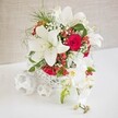 A striking floral centerpiece combines white and red blooms in a composition that feels both contemporary and timeless, perfectly at home on a wedding table or special celebration setting in Grove Park. The arrangement is gathered within a low, round white basket-style container, its open-weave construction forming a delicate lattice that allows glimpses of stems and gives the piece a light, crafted quality. Rising from this base, several large white Oriental lilies open widely, their sculptural petals curving gently back to reveal bold stamens tinged with golden pollen and pale green throats. Intertwined with the lilies are velvety red roses, their petals tightly layered into deep, romantic cups of rich crimson, offering a striking contrast to the cool white blooms. Fine sprays of white baby's breath thread delicately throughout, creating a soft, misty halo around the bolder flowers, while slender, bright green foliage and feathery greenery add freshness and subtle movement, echoing the greenery of local Grove Park gardens. Scattered among the leaves, shiny red hypericum berries form small, rounded clusters that contribute a festive, almost jewel-like accent, perfect for winter celebrations or elegant evening gatherings. On the right side of the display, a graceful spray of white phalaenopsis orchids cascades downward, each flower with smooth white petals and a petite yellow-and-blush centre, with a short line of unopened buds extending towards the tip. On the left, three decorative white woven spheres rest gently on the table, mirroring the container's texture and adding sculptural interest. The whole arrangement radiates refined romance and serenity, creating an inviting focal point for intimate dinners, weddings, or anniversary celebrations across Grove Park and neighbouring riverside areas.