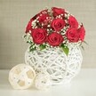 This image presents an intimate, wedding-inspired table arrangement built around a dome of deep red roses, the kind of refined centrepiece that could adorn a reception in Grove Park. At the core is a tightly composed bouquet of roughly a dozen to fifteen roses, each one beautifully open, with velvety petals curling inward in layered spirals, creating a plush, almost sculpted surface. Among the crimson blooms, tiny white flowers of baby's breath are threaded delicately, their airy clusters drifting between petals and adding contrast, lightness, and a feathery texture. Small flashes of green foliage peek from the edges, suggesting the living stems beneath and enhancing the bouquet's natural charm without disturbing its rounded form. The flowers sit in a striking spherical vase, entirely white and shaped from interwoven strands that form an open, lattice-like shell. This woven structure resembles modern wicker or rattan, giving the vase a handcrafted, artisanal character that pairs beautifully with the roses' classic romance. Resting on a smooth white table surface, the vase shares the scene with two decorative orbs to its left, each made of pale, flat woven strips that create open, airy spheres in soft cream tones; one is larger and closer to the front, while the smaller nestles just behind, echoing the vase's pattern. The background is a softly blurred wall in warm beige, with a subtle texture that suggests fabric or finely plastered stone, ensuring the bold reds and crisp whites remain the focus. The overall atmosphere is calm, elegant, and warmly romantic, ideal for illustrating wedding table flowers, romantic dinners, or sophisticated home styling in and around Grove Park.