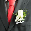 This close-up focuses on a timeless boutonniere pinned to the lapel of a deep charcoal suit, a familiar sight at weddings and proms around Grove Park. At the heart of the piece is a single white rose, luminous and gently opening, each petal layered in a natural spiral that suggests sincerity and new beginnings. The rose's soft, velvety texture contrasts beautifully with the smooth weave of the suit fabric, drawing the eye to the floral detail. Around the base of the bloom, tiny white baby's breath blossoms cluster in delicate sprays, creating a cloudlike collar that adds volume without overpowering the central flower. Slender ferns and smooth, looping greenery stretch slightly beyond, giving the design a touch of movement and a nod to the surrounding greenery found along Grove Park's quiet residential streets. A narrow white satin bow rests at the bottom of the arrangement, tied with precision, its sheen catching the light just enough to add a refined accent. The stems are neatly wrapped and pinned flat, ensuring the boutonniere sits close to the chest, comfortable for a full day of greetings, dancing, and toasts. The overall mood is serene and polished, as though the wearer is taking a final deep breath before stepping out to meet guests. Whether chosen for a groom, best man, or an honoured guest, this floral piece feels perfectly at home in the relaxed elegance of Grove Park and nearby Chiswick, carrying a quiet promise of love, celebration, and cherished memories.
