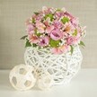 A lush, contemporary floral centerpiece sits on a pristine white tabletop, its rounded form glowing softly against a textured light beige wall that feels like calm afternoon light in a Grove Park townhouse. The white decorative vase is the first thing your fingers would find: a weighty, bulb-shaped container built from slender white strips woven into an open lattice, almost like sculpted wicker or rattan, with gaps that let the background gently show through. From this modern basket-like base rises an abundant bouquet of pastel pink and lavender roses, their velvety petals layered and full, opening outwards in generous spirals that suggest quiet romance and care. Threaded between the roses are graceful pink alstroemeria, or Peruvian lilies, each bloom trumpet-shaped with fine darker pink or soft yellow streaks painted along the inner petals, giving the arrangement a more intricate, almost hand-painted feel. Bright lime green button chrysanthemums add a playful burst of colour and a compact, pom-pom texture, dotting the design like cheerful confetti. Wisps of white gypsophila (baby's breath) float between the larger flowers, cloud-like and airy, softening the edges and giving the whole piece a dreamy, weightless quality. Deep green leaves peek out here and there, grounding the pastel palette with natural depth and structure. To the left of the vase, two decorative woven spheres in light, natural tones sit on the white surface, one slightly larger and tucked closer to the base, the smaller just in front, echoing the open-weave of the vase and tying the composition together. The overall mood is elegant yet relaxed, ideal as a spring or summer centerpiece for a Grove Park dining table, a gentle birthday surprise, or a thoughtfully styled arrangement for a quiet celebration at home.