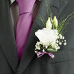 This wedding boutonnière, pinned neatly to a dark suit that could be worn for a ceremony near Grove Park station or a riverside venue by the Thames, offers a beautifully restrained expression of romance. The central focus is a fully open white lisianthus bloom, its petals layered in soft, ruffled whorls that seem to ripple outward from the centre, giving the flower an almost cloud-like fullness. The white is pure but gentle, edged with the faintest shadows in the folds, creating natural depth and texture. Just behind, two slim lisianthus buds stand in quiet poise, their pale green casings wrapped snugly around the petals within, suggesting the unfolding of new chapters in a shared life. Encircling the main flower, clusters of baby's breath scatter in tiny, starry blossoms, creating a light, misty halo that softens the strong line of the suit's lapel. Fine green blades of foliage weave between the blooms, bringing a slight touch of woodland spontaneity, as if the stems were freshly chosen from a local Grove Park garden on a cool morning. At the base, a deep-green, glossy leaf is rolled and curved with intention, forming a natural frame that supports and grounds the composition. A slender mauve ribbon is tied into a precise bow around the stems, the soft lavender hue echoing the groom's tie in a subtle, coordinated gesture, its satin surface catching mild highlights from the surrounding light. The overall mood is one of calm refinement and heartfelt sincerity, as though you could sense a gentle, clean floral scent rising from the boutonnière whenever the groom moves, carrying with it the quiet joy and anticipation that define the wedding day.