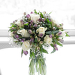 In this thoughtfully composed sympathy bouquet, white and soft purple blooms rise from a clear glass cylinder vase, bathed in bright, diffused light that gives the scene a calm, almost timeless atmosphere, as if set in a quiet Grove Park home. The primary flowers are creamy white roses, their petals smooth and full, opening in gentle spirals that suggest tenderness and quiet devotion. Woven among them are numerous alstroemeria stems, some in tight bud, others opening into elegant, trumpet-like blooms in hues of mauve, lilac, and light violet, with subtle green touches at the petal bases. These colours provide a muted contrast to the roses, adding depth without disturbing the bouquet's peaceful mood. Airy clusters of baby's breath are scattered throughout, their tiny white flowers creating a fine, lace-like texture that softens every edge and adds a sense of lightness, as if the arrangement were wrapped in a gentle, floral haze. Lush green foliage supports the entire composition: broad, glossy leaves and narrower, arching greens form a natural frame, enhancing the brightness of the white roses and the cool purples, much like the greenery surrounding local paths towards the River Thames. The glass vase is simple and transparent, allowing the straight green stems to be seen clearly, standing in clean water and emphasising the bouquet's freshness and sincerity. Against a luminous, almost pure white background, the arrangement stands out with quiet clarity. The overall feeling is one of comfort and reflection-a floral tribute well suited to expressing sympathy, remembrance, and heartfelt support for families and friends throughout Grove Park.
