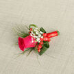 This boutonniere is built around a single, striking fuchsia rose, open enough to reveal its rich, velvety centre yet still holding the gentle curves of a flower in its prime. Surrounding it, tiny white blossoms of baby's breath cluster together, giving a soft, cloud-like frame that contrasts with the saturated colour of the rose. Nestled close to the base, red hypericum berries add rounded shapes and a deeper red tone, offering a playful yet sophisticated accent. The foliage has been folded and looped with care: broad green leaves cradle the flowers in a loose embrace, while slender grass-like strands arc and weave through the design, creating a sense of lightness and flow. A red satin ribbon wraps the stems neatly, its smooth texture and subtle sheen bringing a refined finish suited to formal wear. Against a pale, textured background, the colours appear vivid but controlled, making every element easy to distinguish-the open rose, the tiny gypsophila flowers, and the polished berries. This piece captures the feeling of a special occasion in Grove Park, perhaps a wedding at a local church or a celebration hosted near the quiet, tree-lined avenues. It suggests careful craftsmanship, the kind of floristry where each stem is placed with intention so that the finished boutonniere feels balanced and comfortable to wear. Imagining it pinned to a jacket, you can almost feel the cool petals brushing against fabric and sense the subtle freshness of cut foliage, carrying with it the quiet promise and excitement of a meaningful day.