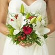 Captured close against a backdrop of fine lace, this hand-tied bridal bouquet glows with understated luxury, the kind of design you might see being carried from a Grove Park florist's studio toward a ceremony near the Thames. At its core is a single, perfectly formed red rose, its velvety petals unfolding in rich layers and drawing the eye into its romantic depth. Around it, creamy white lilies open gracefully, their star-shaped faces and slightly ruffled edges catching the soft light. The lilies' smooth petals contrast beautifully with delicate sprays of gypsophila, whose tiny white blooms gather like a light mist around the central flowers. Clusters of glossy red berries appear throughout, adding playful bursts of colour and a sense of joyful festivity. Between them, moss-green dianthus and textured foliage provide a soft, earthy backdrop, evoking the natural calm of nearby Grove Park and Chiswick's hidden gardens. Slender blades of ornamental grass weave through the bouquet, giving a sense of movement and airiness, while the mix of rounded mossy forms and linear greens adds sculptural interest. The whole arrangement is gathered tightly at the stems and wrapped in smooth ribbon, which sits comfortably in the bride's hands. A few lily buds, still closed and tipped with pale green, peek out from the arrangement, hinting at moments and memories still to unfold in the couple's shared life. The bouquet's blend of white, red, and rich green creates an elegant balance of purity and passion. It feels like a heartfelt expression of devotion and anticipation, captured in flowers and crafted with the quiet confidence of a local Grove Park florist.