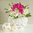 This floral arrangement appears like a still moment from a wedding morning in Grove Park, captured in soft, diffused light. At its heart lies a compact mound of vivid magenta-fuchsia blooms, their petals densely layered and gently ruffled so that they form a lush, cushion-like centre, radiating warmth and romance. Surrounding these central flowers, a halo of creamy ivory alstroemeria unfolds, each blossom composed of slender, elongated petals that curve outward with effortless grace; fine veins of pink and fresh green trace their surfaces, lending an artistic, almost hand-illustrated detail to the design. Slender green stems and a scatter of small buds rise above and between the blossoms, punctuating the arrangement with crisp accents and a sense of life in motion, like new leaves opening along the quiet streets near Grove Park station. The flowers are gathered closely in a low, round basket crafted from white-painted wicker; its intricate weave loops and crosses with a gentle rhythm, providing a clean, modern frame for the vivid colours above. Nearby, two creamy woven orbs rest on the tabletop, echoing the basket's texture and adding sculptural interest that feels minimalist yet inviting. The overall light is cool and gentle, creating soft reflections on the wicker and subtle shadows between petals, which emphasise depth without overwhelming the scene. This bouquet feels serene yet celebratory, perfectly suited for wedding receptions, bridal showers, or registry office gatherings in and around Grove Park, and ideal for same-day delivery to mark engagements, anniversaries, or heartfelt thank you moments with an arrangement that balances romance, freshness, and carefully considered design.