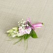 A delicately crafted corsage or boutonniere lies centered on a softly woven, oatmeal-coloured linen background, its neutral fabric texture gently catching the light and giving the whole scene a calm, elegant feel, as if laid out on a dressing table before a Grove Park wedding. At the heart of the piece, a tight cluster of soft pink, multi-petalled blooms-reminiscent of miniature stocks or ruffled garden flowers-unfolds in gentle layers, their petals moving from a paler blush at the edges to a slightly warmer pink at the centre. Threaded between these are tiny white clouds of baby's breath, each floret no bigger than a pinhead, creating a light, lacy halo that softens the structure. Here and there, subtle accents of lavender and deep purple flowers add quiet depth, like little twilight notes in an otherwise pastel melody. On one side, creamy white to pale green buds, likely freesias, stretch outward in a graceful line, their slender forms suggesting blooms about to open, full of quiet promise. The arrangement is cradled in glossy, broad green leaves that fan gently beneath, with fine, feathery fronds of grass-like foliage escaping at the edges to lend an effortless, garden-picked charm you might associate with walks through nearby Grove Park or along the tree-lined streets off Burlington Lane. The stems are neatly gathered and bound in a vivid fuchsia satin ribbon, tied into a perfectly balanced bow whose smooth loops and short trailing ends add a joyful, celebratory touch. The soft, even lighting highlights every fresh petal and leaf, creating an intimate portrait of a handmade floral accessory ready for a wedding, prom, or special event in West London.