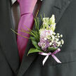 Pinned carefully to the lapel of a dark, well-tailored suit, this boutonnière appears as a small, exquisite garden composed on the most miniature of canvases. At its heart, soft lilac flowers unfold in delicate layers, their petals curling slightly at the edges in a way that suggests both tenderness and strength, like the early spring blossoms you might spot in front gardens along Grove Park Road. Creamy, unopened buds nestle alongside them, promising bloom and gently echoing the softness of the wearer's shirt. Around this central cluster, graceful, curved green leaves create a smooth, glossy frame, their surfaces catching the light with a subtle shine that hints at dewy freshness. Thin wisps of feathery greenery rise and arch outward, adding a sense of light movement and a touch of wildness, as though the boutonnière had just been gathered from a hedgerow near Chinbrook Meadows. Sprays of tiny white gypsophila weave between the petals, forming a fine, airy texture that softens every edge. The stems are bound tightly at the base with a lilac ribbon, its sheer, satiny sheen mirroring the nearby tie and bringing all the colours into gentle harmony. The overall impression is one of quiet romance and considered style, the kind of floral detail a local Grove Park florist would design for an intimate wedding, civil ceremony, or special family occasion, where each bloom is chosen to tell a subtle story of love, care, and celebration.