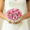 In this photograph, the focus rests on a romantic bridal bouquet held close to an ivory lace dress, capturing a quiet moment before a Grove Park wedding. The bouquet is a lush, rounded cluster of blush pink roses, each rose fully open, with layers of satiny petals forming gentle, spiralling cups. The colour transitions subtly from a warm, rosy blush at the heart of each bloom to paler, almost creamy edges, creating depth and softness all at once. Interwoven between the roses is a generous scattering of white gypsophila, its tiny blossoms clustered on fine stems like delicate stars, giving the arrangement a light, airy halo. The gypsophila softens the outline of the bouquet and mirrors the intricate, lace-like detail of the bride's gown, tying the whole composition together visually. Soft, natural light seems to fall from one side, suggesting an indoor setting near a bright window in a Grove Park venue or family home, with the light gently illuminating the bouquet without harsh shadows. The bride's hands are relaxed yet secure around the stems, as though pausing for a deep breath before stepping out into a garden or onto a leafy Grove Park street lined with friends and family. The overall impression is one of gentle romance and calm anticipation, as if the bouquet quietly holds the emotions of the day-love, hope, and lasting commitment. You can almost imagine the scent of the fresh roses, lightly perfumed and uplifting, drifting through the room and mingling with the soft murmurs of guests gathering just outside.