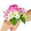 A delicate wrist corsage rests against a clean white backdrop, shown on a left hand and forearm with a light-to-medium skin tone, the composition lit softly as if by morning light. At the centre sits a generous, fully opened deep fuchsia-pink rose whose tightly layered petals spiral outward with velvety texture; its scent seems almost visible, a warm, sweet note that anchors the piece. Surrounding the rose are clusters of blush-pink hydrangea florets, their papery, cloud-like petals building a soft, voluminous base that cushions the focal bloom. Scattered between them are several white alstroemeria (Peruvian lilies), each with four to six slender petals and pale green streaking at the throat, adding a crisp, refined contrast to the richer pinks. To the right a small cluster of pale green alstroemeria buds and a few barely opened pink buds suggest promise and movement, a floral heartbeat hinting at more to come. Broad, glossy emerald-green leaves frame and lift the arrangement, their smooth surfaces catching light and giving depth and contrast to the pastel palette. The entire ensemble is fastened to the wrist by a refined multi-strand bracelet of small, luminous white pearls that wraps neatly around the wrist and trails slightly down the back of the hand, lending a timeless, wearable elegance. This corsage evokes quiet celebration-prom night, a wedding vow, or an afternoon at Chinbrook Meadows-with craftsmanship reminiscent of local Grove Park floristry and a tactile beauty that invites a gentle touch.