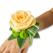A delicate wrist corsage rests gently on the inner wrist, its warm tones and crisp greens captured against a clean white backdrop. At the centre blooms a single, fully opened peach-apricot rose, its velvety petals unfurling in soft spirals that catch a quiet, morning light; the rose reads as tender and reassuring, fragrant with the faintest whisper of classic tea-rose perfume. Tucked to the lower left of the bloom are two lively lime-green button chrysanthemums-small, spherical pom-poms whose tightly packed florets add a playful, textured contrast like bright notes in a soft melody. The flowers are cradled on a bed of deep green foliage: broad, glossy leaves-likely ruscus or salal-provide a lush foundation, while a long, variegated Ti-style leaf with creamy horizontal stripes is artfully curled and folded, sweeping from beneath the rose toward the outer edge of the wrist and lending a modern, architectural flourish. The corsage appears secured by an invisible band beneath the leaves, so the arrangement seems to float atop the skin. The overall palette of peach, lime and rich greens feels quietly celebratory, suited to a wedding morning, prom or intimate anniversary; it radiates a gentle anticipation and would pair beautifully with soft champagne or ivory attire. As a local florist might note, the piece is handcrafted with attention to balance and movement-soft in scent, varied in texture, and reassuring in scale-an elegant wristlet designed to be both worn and remembered in the small, tender moments of a special day.