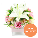 Captured against a bright, uncluttered white background, this floral arrangement radiates a quiet elegance that echoes the calm streets and riverside walks of Grove Park. Two fully opened white Oriental lilies form the central focal point, their large, gently curving petals spreading out like stars and revealing tall, reddish-brown stamens at their centres. Surrounding these lilies is a cushion of pink roses, each at a different moment of bloom-from tightly furled buds that hint at opening to fully expanded flowers with many soft, velvety petals layered in romantic spirals. Threaded between the roses and lilies, alstroemeria blossoms in shades of pink and white add a graceful, slightly wild note. Their delicate petals are marked with fine yellow strokes and subtle speckles that introduce a sense of complexity and depth. Fine sprays of white baby's breath drift throughout the bouquet, creating a light, cloud-like softness and gently filling in the spaces between the larger blossoms. A variety of green foliage frames the display: some leaves are broad and glossy, catching the light in smooth planes, while other stems carry finer, feathery fronds that bring gentle movement, recalling leafy corners of Dukes Meadows. All the stems are anchored neatly in a contemporary square glass vase, crystal clear so that the lower part of the stems and the clean water are visible. Around the middle of the vase, a pale pink satin ribbon is tied in a simple horizontal band, echoing the pastel tones of the roses and giving the piece a polished, gift-ready feel. In the bottom right corner of the image, a bright orange circular graphic with the words VASE INCLUDED in bold white capitals signals clearly that the vase is part of the arrangement. The scene feels serene and thoughtful, perfect for a modest dining table in Grove Park or as a gentle, heartfelt delivery for a friend or loved one.